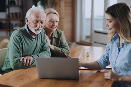 Image of a young woman sitting across a table from an elderly man and woman who are learning into each other. All are looking at a laptop that the young woman is pointing to.