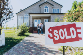 Image of a sold sticker on a for sale sign in the front yard of a house with a family standing in front of it.