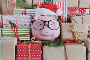 Image of a ceramic piggy bank wearing glasses and a Santa hat sitting on a stack of presents.